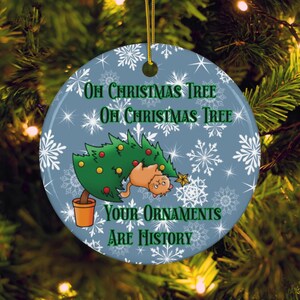 May include: A round Christmas ornament with a light blue background and white snowflakes. The ornament features a green Christmas tree with a cat and the text "Oh Christmas Tree, Oh Christmas Tree, Your Ornaments Are History."