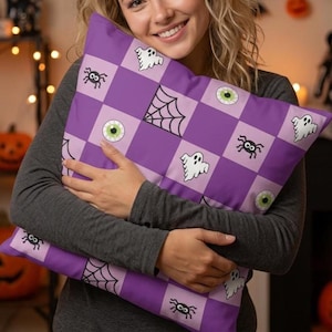 May include: A square throw pillow featuring a purple and light purple checkerboard pattern. The design includes repeating black and white illustrations of ghosts, spiders, spiderwebs, and green eyeballs distributed across the alternating squares.