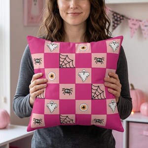 May include: A square throw pillow featuring a checkered pattern of alternating hot pink and light pink squares. The design includes repeating black and white illustrations of ghosts, spider webs, spiders, and realistic human eyeballs.