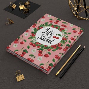 May include: A pink journal with a cherry pattern and the words "Life is Sweet" on the cover. The journal is surrounded by gold and black pencils, paper clips, and a decorative gold geometric object.