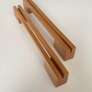 Könnte beinhalten: Zwei hellbraune Holzständer, die jeweils mit einem langen, schmalen vertikalen Schlitz an der Oberseite versehen sind. Diese rechteckigen Holzhalter haben eine glatte Oberfläche und sind parallel zueinander auf einer schlichten Fläche angeordnet.