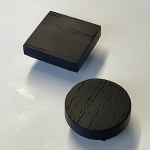 Può includere: Due oggetti in legno neri: un quadrato e un cerchio. Il quadrato è leggermente rialzato. Entrambi hanno un aspetto di venatura del legno strutturata. Il cerchio è in basso e il quadrato in alto.