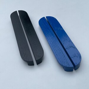 Può includere: Due supporti per telefono in legno, uno nero e uno blu, sono esposti su uno sfondo bianco. Ogni supporto ha una parte superiore curva e una fessura per tenere un telefono. I supporti sono in legno.