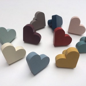 Può includere: Una collezione di oggetti in legno a forma di cuore in vari colori pastello, tra cui blu, rosa, giallo e grigio. Gli oggetti sono disposti su una superficie bianca, mostrando la loro forma tridimensionale e la finitura liscia.