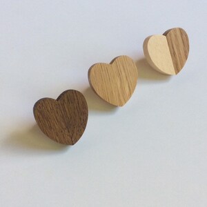 May include: Three wooden heart-shaped objects in shades of brown and tan are arranged diagonally on a white surface. The hearts have a natural wood grain texture, with one featuring a two-tone design. These appear to be decorative or craft items.