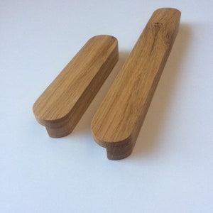 May include: Two wooden cabinet pulls with a natural wood grain finish. The pulls are rectangular with rounded ends and a small, curved base. One pull is slightly longer than the other. The pulls are made of solid wood and are likely for furniture.