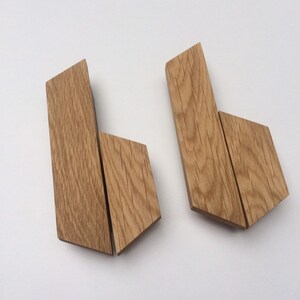 Può includere: Due ganci da parete in legno dal design geometrico. Ogni gancio è realizzato in legno marrone chiaro e presenta una forma rettangolare con bordi angolati. I ganci sono esposti su uno sfondo bianco.