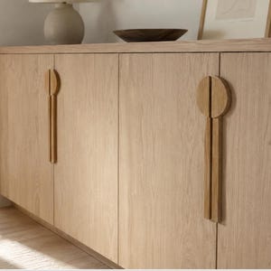 May include: A light oak wood sideboard featuring four cabinet doors with a visible wood grain pattern. Each door is equipped with a unique vertical handle consisting of a circular top section and a long, rectangular lower extension in matching wood.
