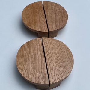 Può includere: Due pomelli per mobili in legno. Ogni pomello è di forma circolare, marrone chiaro, diviso in due metà con uno spazio verticale. I pomelli hanno una base corta e rettangolare. La venatura del legno è visibile.