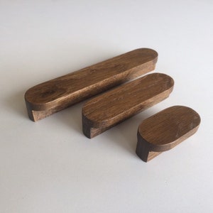 Könnte beinhalten: Drei Holzmöbelgriffe in verschiedenen Größen. Die Griffe sind warmbraun mit glatter Oberfläche und abgerundeten Kanten. Der kleinste Griff ist etwa 5 cm lang, der größte etwa 15 cm.