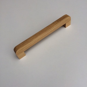 Pode incluir: Um puxador de armário retangular de madeira clara, com uma extremidade arredondada e uma extremidade quadrada. O veio da madeira é visível. O puxador tem aproximadamente 15 cm de comprimento.