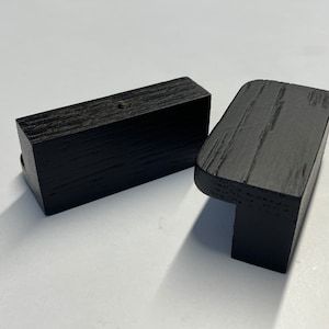 Può includere: Due pomelli per mobili in legno nero. Uno è rettangolare, l'altro ha un bordo arrotondato. I pomelli hanno una finitura scura e strutturata, probabilmente per mobili. Sono mostrati su sfondo bianco.