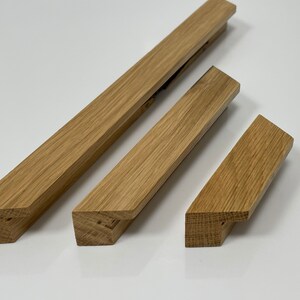 Op de afbeelding: Drie houten fotolijsthangers in verschillende maten. De hangers zijn gemaakt van lichtgekleurd eikenhout en hebben een rechthoekige vorm met schuine randen. De houtnerf is zichtbaar. De hangers zijn gerangschikt op een wit oppervlak.