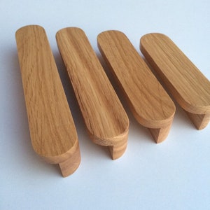 Pode incluir: Quatro puxadores de armário de madeira com acabamento em carvalho natural. Cada puxador tem um topo retangular com extremidades arredondadas e uma pequena base angular. Os puxadores estão dispostos em uma fileira contra um fundo branco.