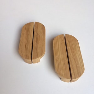 Puede incluir: Dos tiradores de armario de madera con un acabado de veta natural. Cada tirador se compone de dos piezas rectangulares redondeadas conectadas, con una divisi&oacute;n vertical en el medio. Los tiradores son de color marr&oacute;n claro y est&aacute;n sobre una superficie blanca.