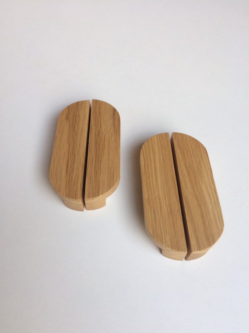 Puede incluir: Dos tiradores de armario de madera con un acabado de veta natural. Cada tirador se compone de dos piezas rectangulares redondeadas conectadas, con una divisi&oacute;n vertical en el medio. Los tiradores son de color marr&oacute;n claro y est&aacute;n sobre una superficie blanca.