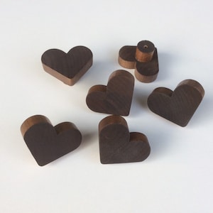 Può includere: Una collezione di oggetti a forma di cuore in legno marrone scuro. I cuori hanno un design bicolore, con un bordo marrone più chiaro e una parte anteriore marrone più scura. Alcuni cuori sono in posizione verticale, altri sono piatti.