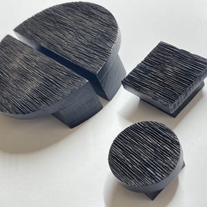 Può includere: Un set di quattro oggetti decorativi in legno nero strutturato. Il set comprende un grande pezzo semicerchio diviso in due, un blocco quadrato e un piccolo pezzo rotondo. Gli oggetti hanno una texture ruvida e lineare.