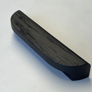 Peut inclure: Objet rectangulaire en bois noir avec un dessus incurvé et une petite encoche à une extrémité. Le grain du bois est visible. Il semble s'agir d'une poignée ou d'un élément décoratif.