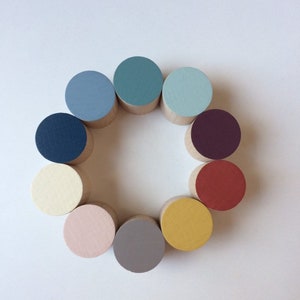 May include: A circular arrangement of twelve wooden blocks, each painted a different colour. The colours include shades of blue, green, red, yellow, and pink. The blocks are arranged on a white background.