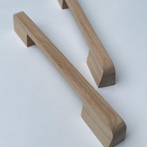 Può includere: Due maniglie per mobili in legno con finitura naturale. Ogni maniglia ha un'impugnatura rettangolare e un manico leggermente angolato. La venatura del legno è visibile e le maniglie sono su uno sfondo bianco.