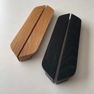 May include: Two wooden phone stands. One is natural oak, the other is black. Each stand has a slot for a phone and a curved top. The stands are on a white surface.
