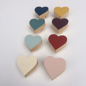 Puede incluir: Una colección de ocho objetos decorativos de madera en forma de corazón. Cada corazón está pintado de un color diferente, incluyendo azul marino, amarillo, verde azulado, burdeos, azul claro, rojo, crema y rosa. Los corazones están dispuestos sobre una superficie blanca.