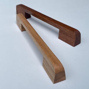 Può includere: Due maniglie per mobili in legno. Una è di colore rovere chiaro, l'altra marrone scuro. Entrambe hanno un design rettangolare con una maniglia curva e sono poste su uno sfondo bianco. Queste maniglie sono ideali per mobili da cucina o bagno.