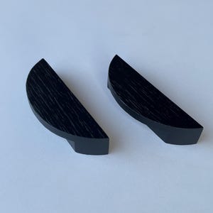 Könnte beinhalten: Zwei schwarze, halbkreisförmige Holzohrringe mit strukturierter Maserung. Jeder Ohrring hat eine kleine, rechteckige Basis. Die Ohrringe sind auf weißem Hintergrund dargestellt.