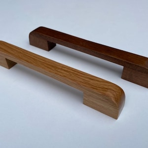 May include: Two wooden cabinet pulls. One is light brown with visible wood grain, the other is dark brown. Both have a rectangular shape with rounded edges and a handle that extends from the base.