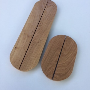May include: Two wooden oval place card holders. One is elongated, the other is smaller. Both are made of light brown wood with a vertical slit down the centre. The holders are designed to hold place cards or small signs.