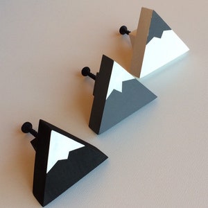 Op de afbeelding: Drie bergvormige kastknoppen. Elke knop is een driehoekig prisma met een zwarte schroef en een zwart, wit en grijs bergontwerp. De knoppen zijn diagonaal op een lichtgekleurd oppervlak geplaatst.