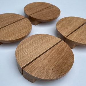 Può includere: Quattro sottobicchieri in legno, ciascuno diviso in due semicerchi. I sottobicchieri sono in legno marrone chiaro con venature visibili e poggiano su piccole basi rettangolari. Il design è semplice e moderno.