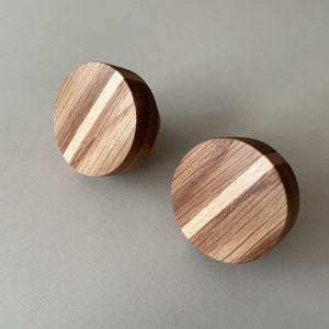 Op de afbeelding: Twee ronde houten kastknoppen. Elke knop is gemaakt van licht- en donkerbruin hout met een diagonale lichtgekleurde streep. Ze liggen op een lichtgrijze achtergrond.