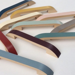 Op de afbeelding: Een verzameling houten meubelgrepen in verschillende kleuren, waaronder marineblauw, geel, roze en grijs. Elke greep heeft een gebogen ontwerp met een geverfde bovenkant en een natuurlijke houten basis. Deze decoratieve grepen zijn ideaal om kleur aan meubels toe te voegen.