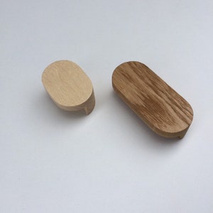 Op de afbeelding: Twee houten kastknoppen. Eén is licht, van nature houtkleurig, en de andere is donkerder, gebeitst. Beide hebben een ovale vorm met een kleine, rechthoekige basis. Deze knoppen zijn waarschijnlijk voor meubels.