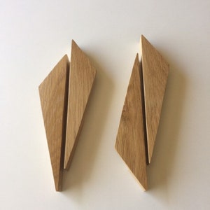 Può includere: Due maniglie per mobili in legno dal design geometrico. Ogni maniglia è composta da tre pezzi angolati di legno chiaro, creando una forma a diamante. Le maniglie sono su uno sfondo bianco.