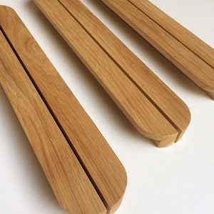 Puede incluir: Tres sujetalibros de madera marr&oacute;n claro, con forma curvada y una ranura central. Los sujetalibros tienen un acabado liso y est&aacute;n dispuestos en diagonal sobre una superficie blanca.