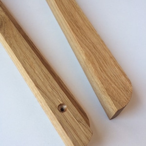 Puede incluir: Dos barras de madera de color marr&oacute;n claro, una con un agujero perforado. Las barras tienen un borde biselado y un extremo redondeado. La veta de la madera es visible. Probablemente son componentes de un art&iacute;culo m&aacute;s grande.