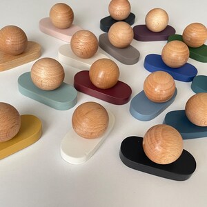 Può includere: Una collezione di pomelli in legno, ciascuno caratterizzato da una parte superiore sferica in legno marrone chiaro naturale fissata su una base piatta di forma ovale. Le basi sono dipinte in vari colori solidi, tra cui bianco, nero, giallo, blu, verde e bordeaux.