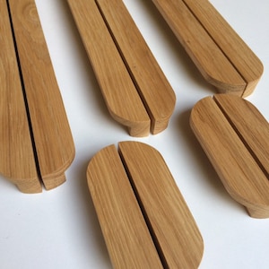 Puede incluir: Varios soportes de tel&eacute;fono de madera clara se muestran sobre una superficie blanca. Cada soporte, de forma oblonga y redondeada, tiene una ranura central y una base plana. Destaca la veta de la madera.