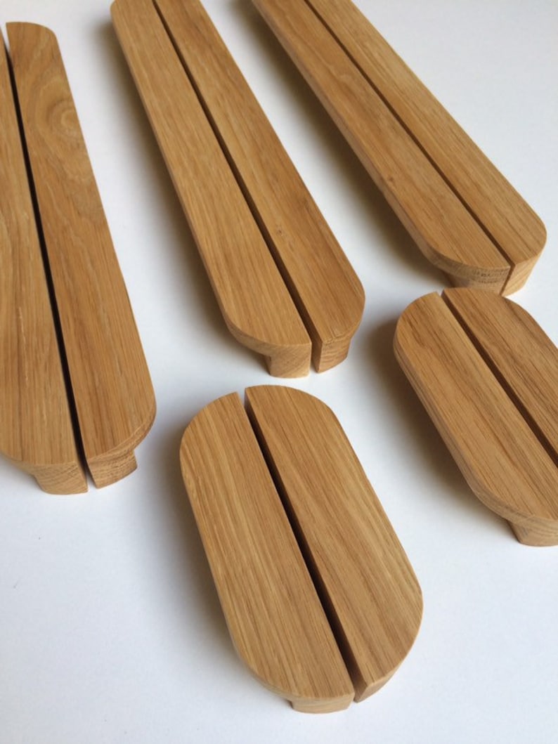 Puede incluir: Varios soportes de tel&eacute;fono de madera clara se muestran sobre una superficie blanca. Cada soporte, de forma oblonga y redondeada, tiene una ranura central y una base plana. Destaca la veta de la madera.