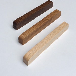 May include: Three rectangular wooden bars in varying shades of brown, from dark to light. The bars have rounded ends and are arranged diagonally against a white background. The wood grain is visible.