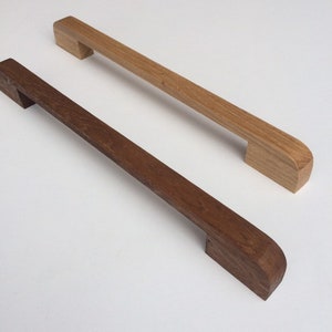 Op de afbeelding: Twee houten meubelgrepen. Eén is lichtbruin, de andere is donkerbruin. Beide hebben een rechthoekige vorm met een gebogen uiteinde. De grepen zijn gemaakt van massief hout en zijn ontworpen voor laden of kasten.