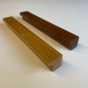 Può includere: Due maniglie per mobili in legno. Una è di colore rovere chiaro, l'altra di colore noce scuro. Entrambe sono rettangolari con un piccolo bordo angolato su un'estremità. Le maniglie sono lunghe circa 25 cm.