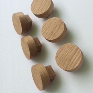 Puede incluir: Seis pomos de madera para muebles. Los pomos son de madera clara y tienen una parte superior redonda y plana con una base cuadrada. La veta de la madera es visible en la superficie. Est&aacute;n dispuestos sobre un fondo blanco.
