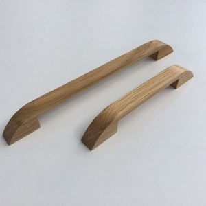 Può includere: Due maniglie in legno per mobili. Una è più lunga, circa 30 cm, l'altra più corta, circa 15 cm. Entrambe sono in legno chiaro con finitura naturale, con un design curvo.