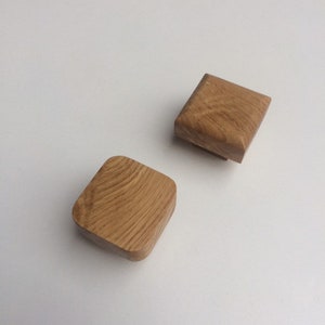 May include: Two square wooden cabinet knobs with rounded corners. The knobs are made of light brown wood and have a visible wood grain pattern. They are set against a white background.