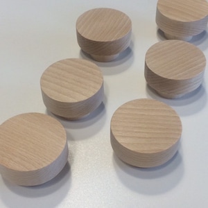 Può includere: Una collezione di sette pomelli in legno non rifiniti, di colore chiaro. Ogni pomello ha una parte superiore rotonda e piatta e una base cilindrica. La venatura del legno è visibile sulle parti superiori. I pomelli sono disposti su una superficie bianca.