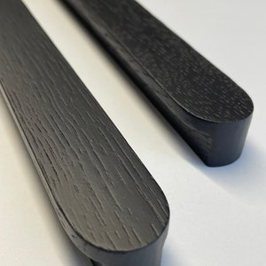 Peut inclure: Deux poignées de meubles en bois noir avec une finition grain de bois texturée. Les poignées ont une forme rectangulaire avec des extrémités arrondies. Les poignées sont présentées sur un fond blanc.